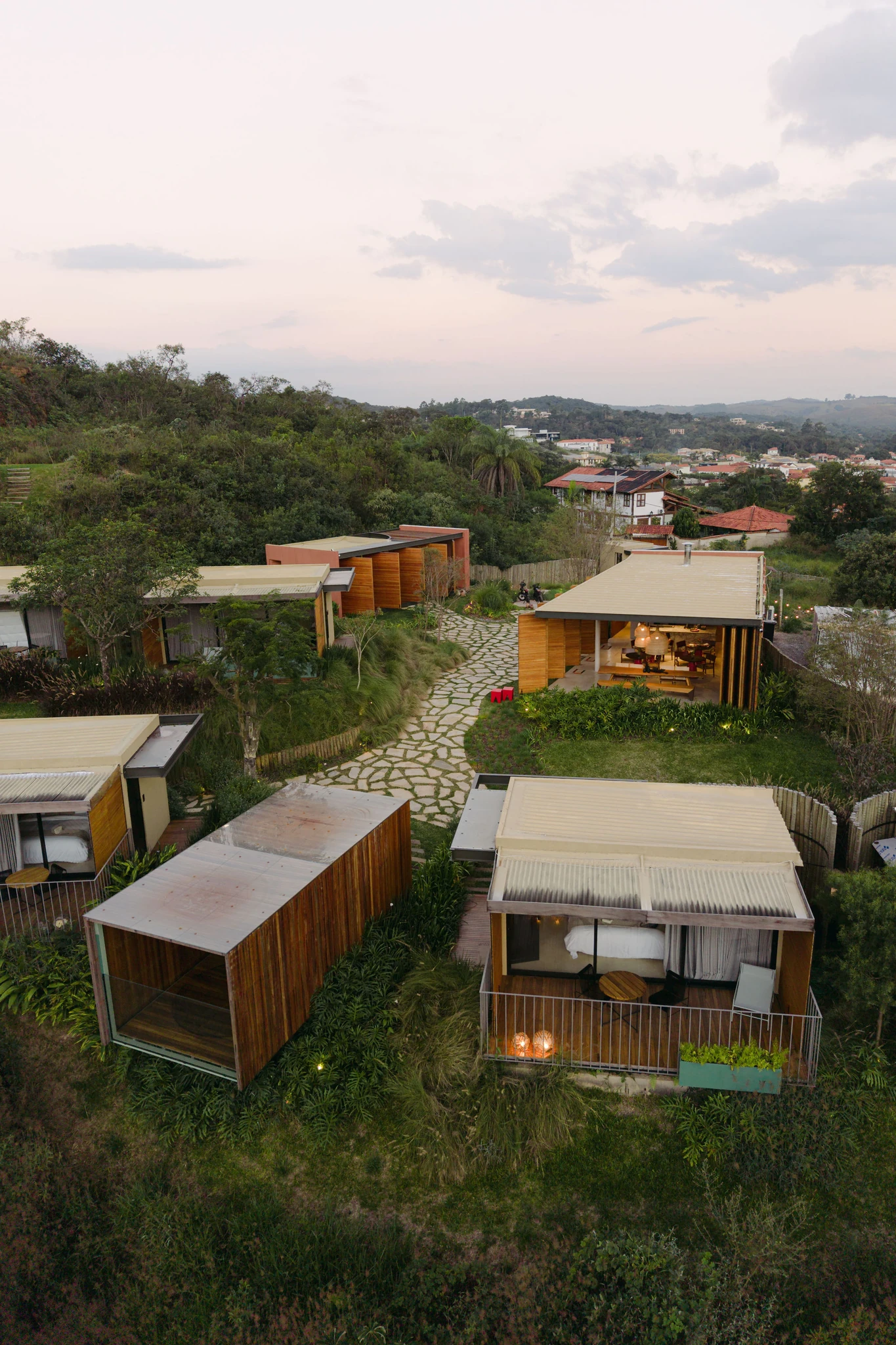Vista aérea do refúgio ao entardecer — suítes, jardim e caminho de pedras, com Tiradentes ao fundo